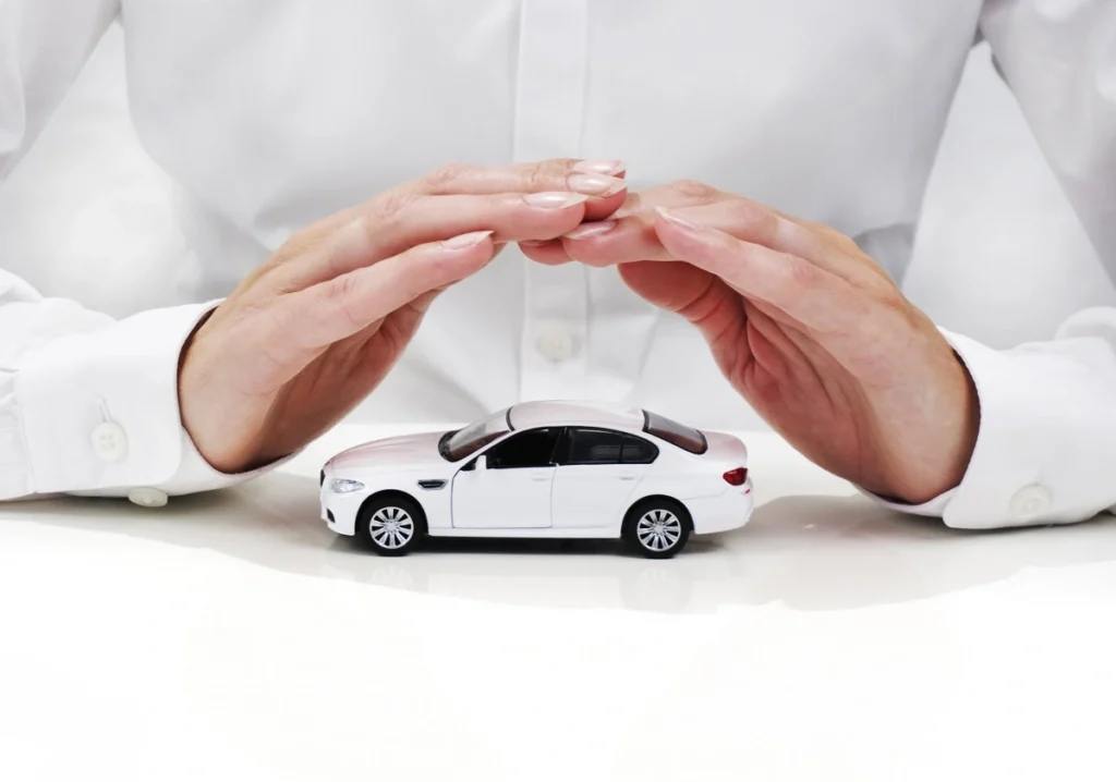 Pessoa faz sinal de proteção com as mãos sobre miniatura de carro na cor branca. Explicando como economizar no seguro auto.