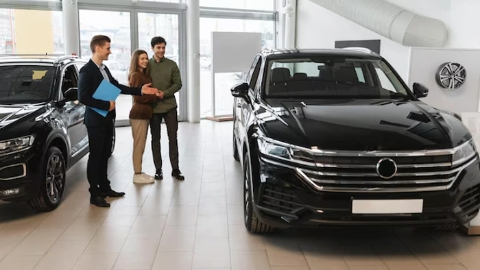 Casal de compradores escuta vendedor de carros em salão. O veículo também está na cena. Falando sobre os melhores seguros para carros.