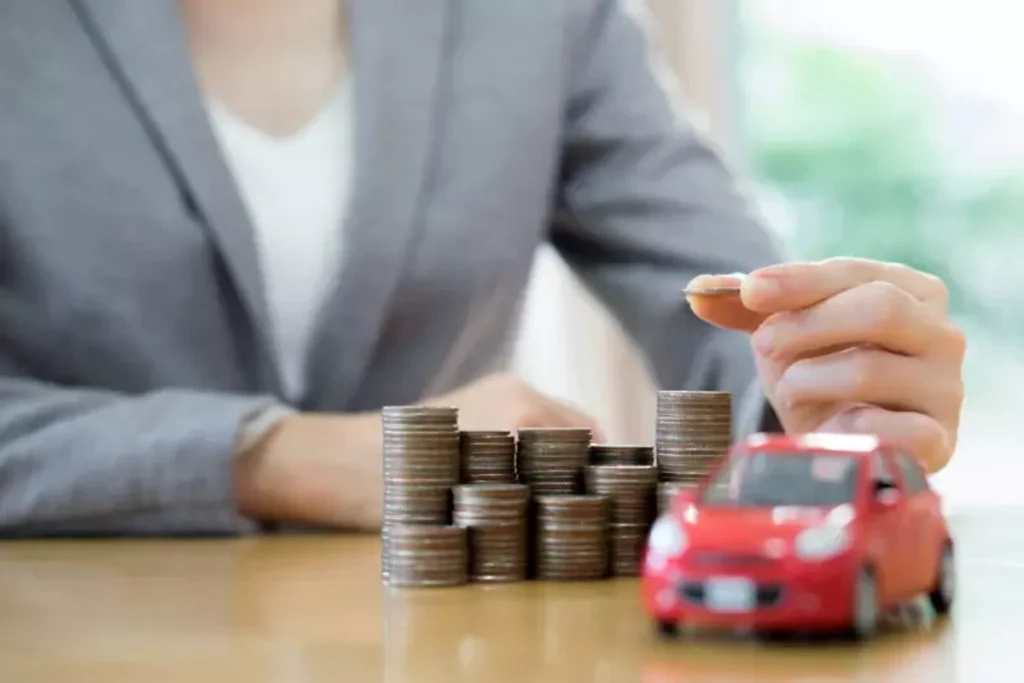 Pessoa coloca moeda em pilha. Há uma miniatura de carro na cor vermelha em cima de uma mesa. Falando sobre como funcionam os consórcios de carros.