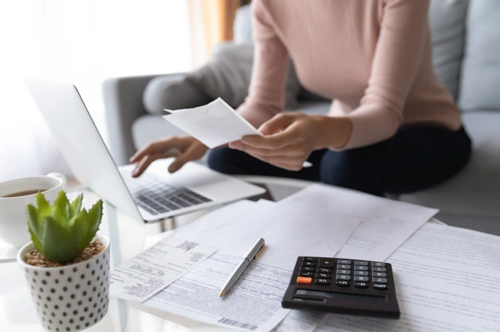 Mulher faz contas com documento usando um notebook e uma calculadora. Há papéis sobre a mesa. Falando sobre qual a diferença de consórcio para financiamento.
