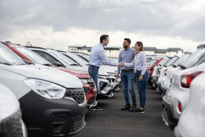Vendedor de carros cumprimenta casal que foi comprar um veículo. Eles estão cercados por vários carros em um pátio. Falando sobre tabela de consórcio de carros.