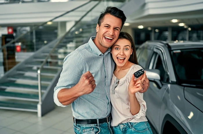 Casal se abraça e sorri dentro de uma concessionária. Atrás deles há um veículo e a mulher está com uma chave de carro nas mãos. Falando sobre como funcionam os consórcios de carros.