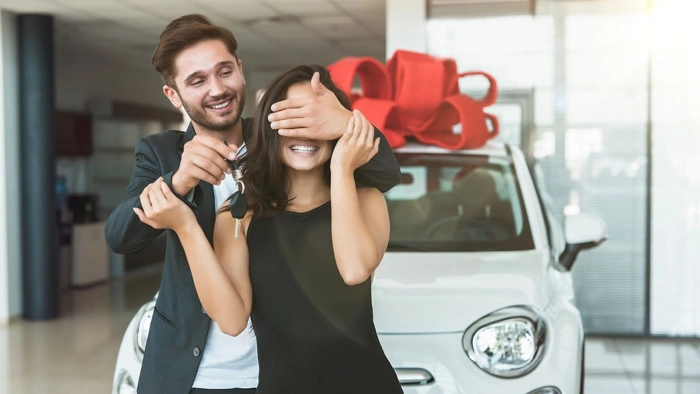 Homem faz surpresa à esposa. Ele tapa os olhos dela com as mãos enquanto carro que será dado de presente está atrás deles, com um laço sobre o teto. Falando sobre consórcio ou financiamento de veículo.
