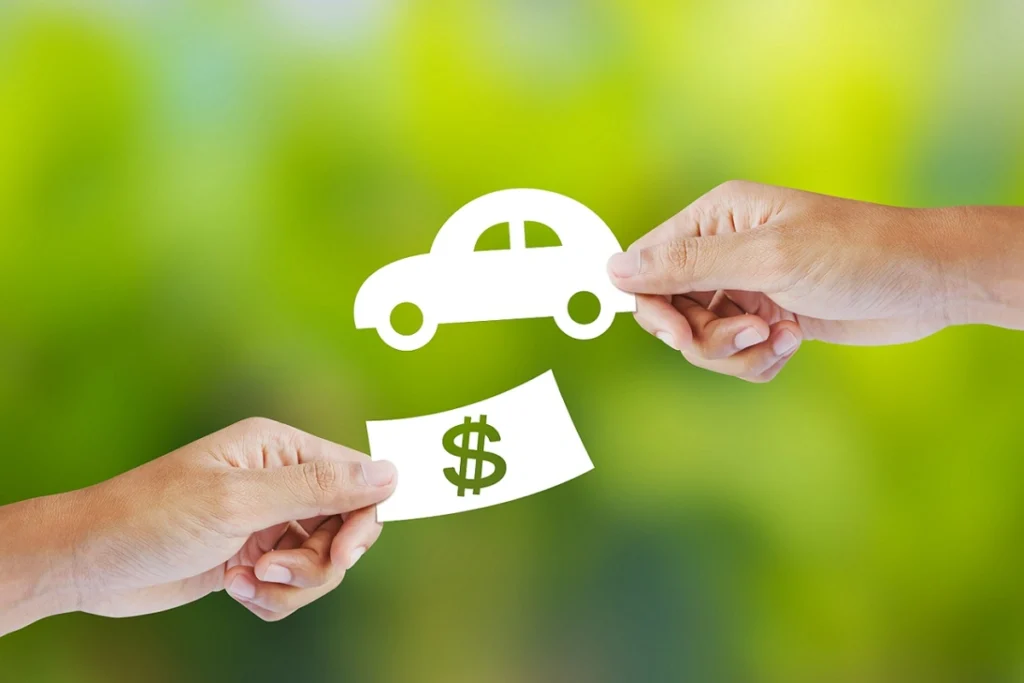 Duas mãos se cruzam sobre um fundo verde. Uma segura uma figura de carro em miniatura e outra de uma figura de uma cédula. Falando sobre consórcio ou financiamento de veículo.