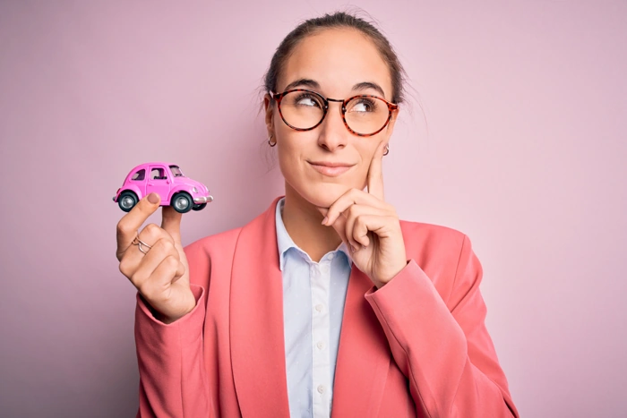 Mulher faz expressão de interesse enquanto segura carro em miniatura na cor rosa com uma das mãos. Falando sobre consórcio para cnpj.